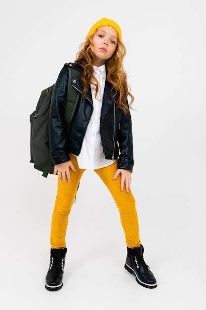 stylish teenager girl in a white shirt, leather black jacket, yellow trousers and a yellow hat, with a backpack on his shoulders posing on white