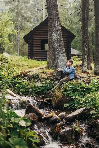 Photo stylish traveler man sitting near cottage and river in sunny forest and mountains travel and wanderlust concept space for text happy hipster guy relaxing in woods summer vacation