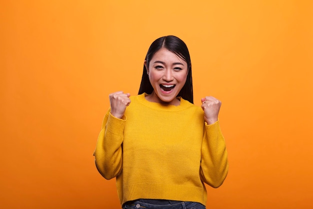 Stylish trendy happy girl celebrating victory and feeling optimistic and independent. Attractive smiling woman raising arms while feeling strong and confident on orange background.