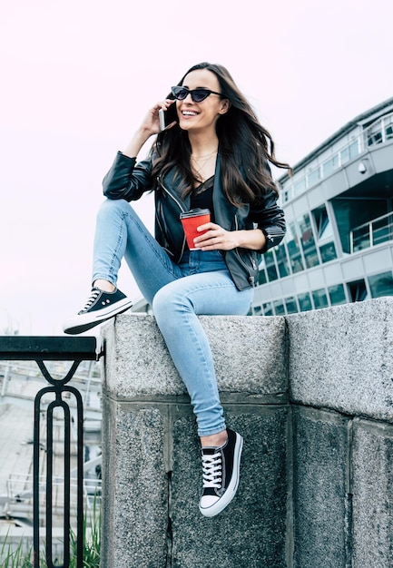 Stylish and trendy young beautiful woman in sunglasses and in hipster style wear while she is using a phone and have a fun outdoors in city