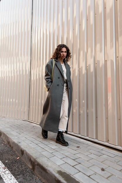 Stylish urban hipster girl with curly hair in a fashionable long green coat with a bag and boots walks on the street near a metal wall