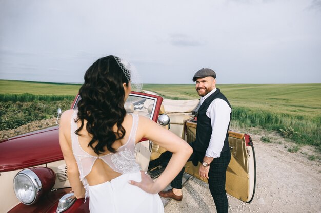 Coppie di cerimonia nuziale alla moda in un campo verde vicino alla retro automobile.