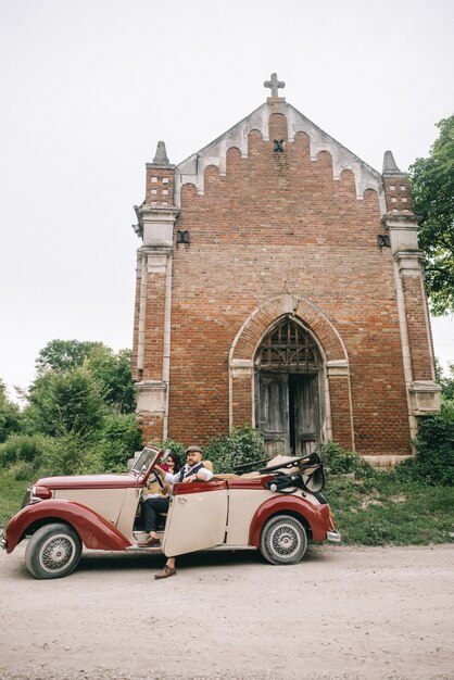 レトロな車で教会の近くのスタイリッシュな結婚式のカップル。