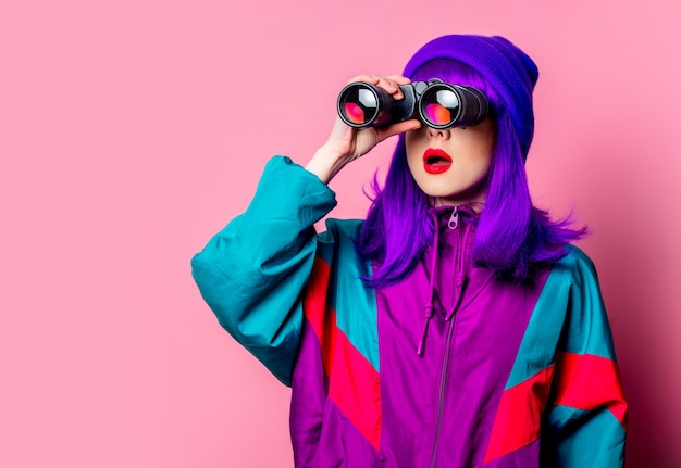 Stylish white girl with purple hair and  tracksuit using binoculars on pink wall