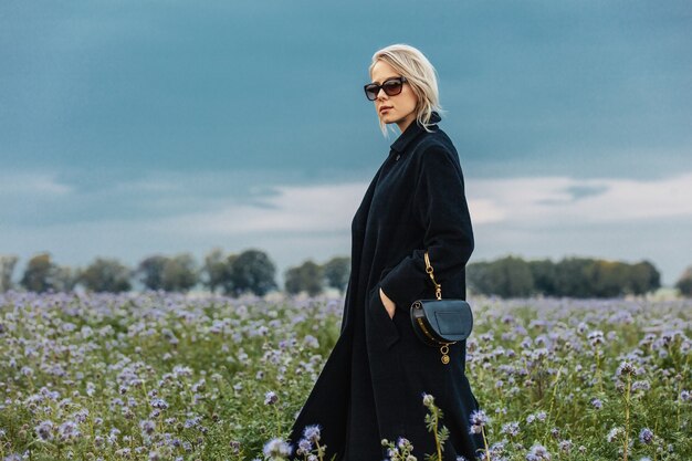 Donna alla moda in cappotto nero e occhiali sul prato con fiori