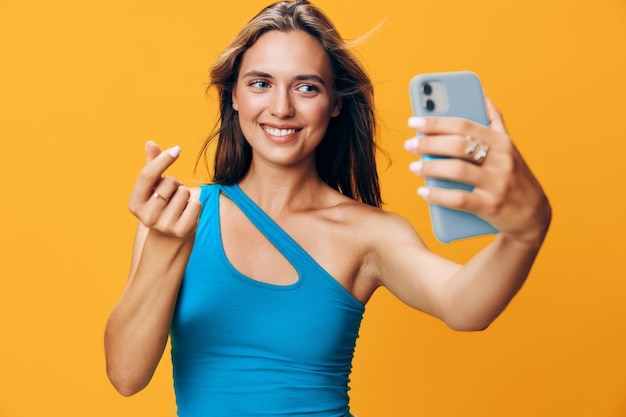 青いトップスを着たスタイリッシュな女性が鮮やかなオレンジ色の背景でスマートフォンで幸せにセルフィーを撮っています