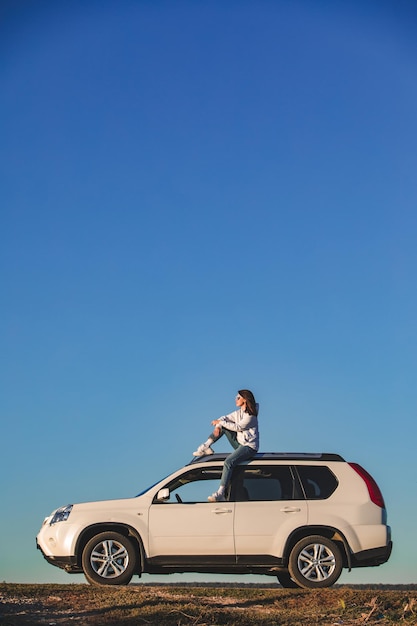 日没時に車の屋根に座っているカジュアルな服装のスタイリッシュな女性