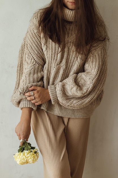 Photo stylish woman in cozy knitted sweater posing with chrysanthemum in hand close up on background of rural authentic wall moody fall cozy home fashionable female with flower autumn aesthetics