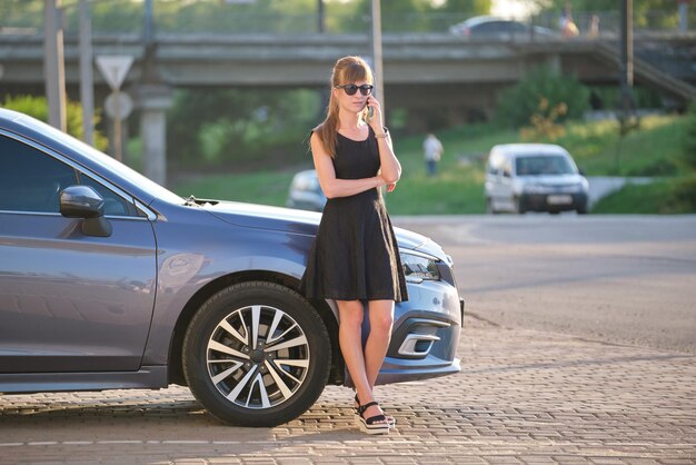 Stylish woman driver standing near her vehicle talking on cellphone on city street in summer