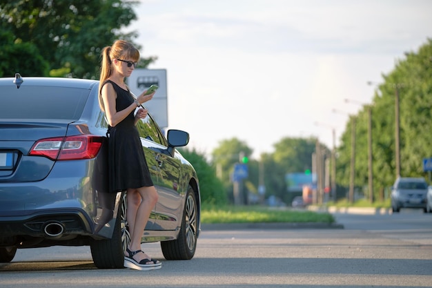 Stylish woman driver standing near her vehicle talking on cellphone on city street in summer