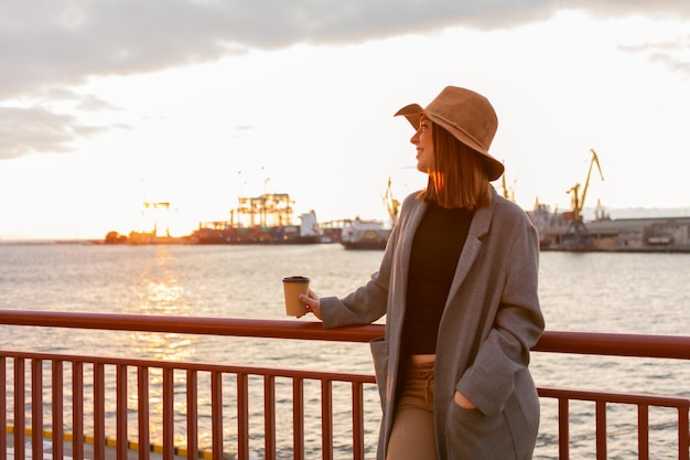 フェルトの帽子とコートを着たスタイリッシュな女性は、海の日の出で外出先でコーヒーを飲みます