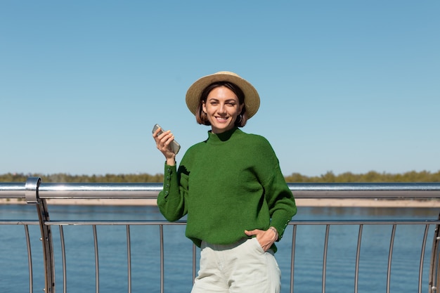 Stylish woman in green casual sweater and hat outdoor on bridge with river view at warm sunny summer day hold mobile phone smile