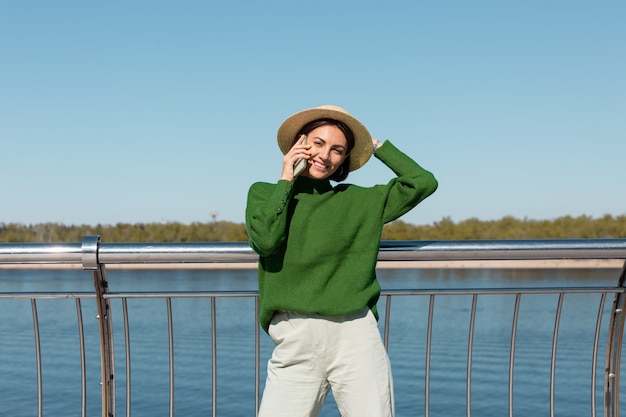 Stylish woman in green casual sweater and hat outdoor on bridge with river view at warm sunny summer day talks on mobile phone