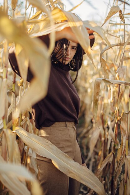 Stylish woman in hat and brown outfit posing in autumn maize field in warm sunny light Fashionable