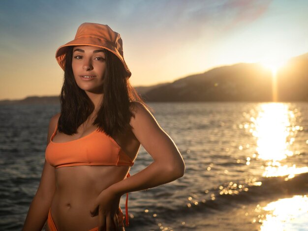 Stylish woman in hat standing near water at sunset