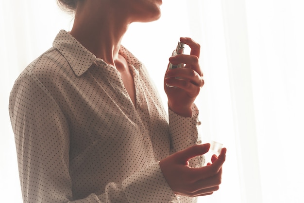 お気に入りの香水のボトルを吹きかけるブラウスのスタイリッシュな女性が自宅でクローズアップ。
