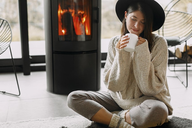 Photo stylish woman in knitted sweater and hat holding warm cup of tea and relaxing at modern fireplace