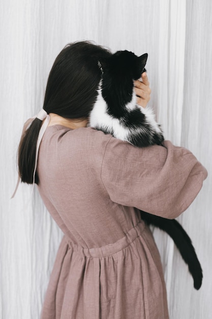 Stylish woman in linen dress hugging cute cat on background of\
pastel fabric simple slow living