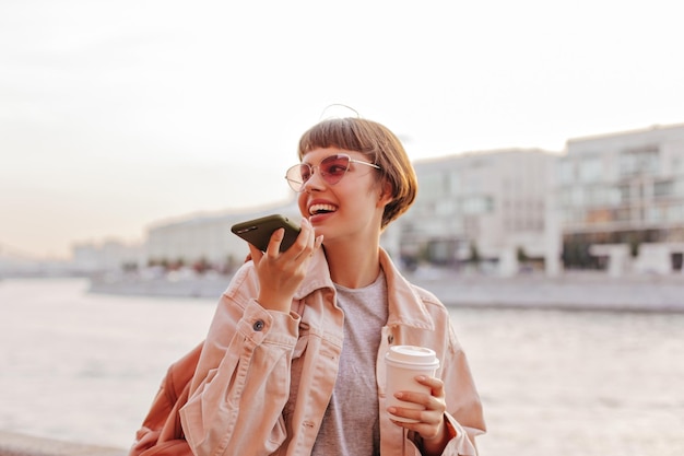 Donna elegante che posa con il telefono in città ragazza adolescente dai capelli corti con occhiali rosa e giacca beige sorride e tiene una tazza di caffè all'aperto