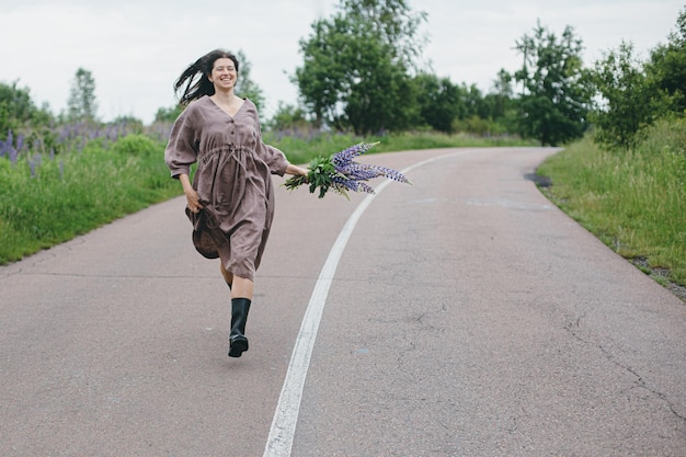 Stylish woman in rustic dress running with lupine bouquet in\
summer countryside cottagecore