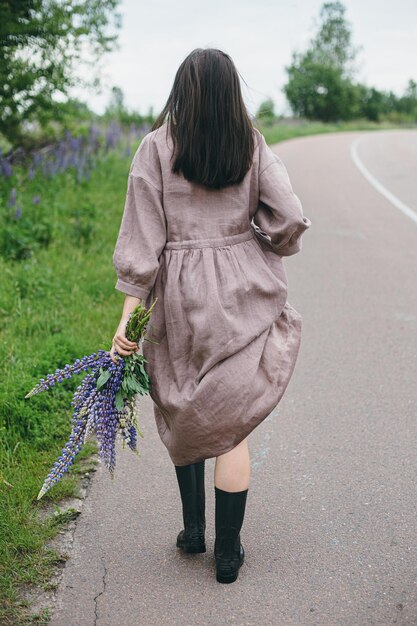 Stylish woman in rustic dress walking with lupine bouquet in\
summer countryside cottagecore aesthetics