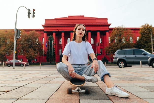 Stylish woman sitting on a longboard on a street background and posing for the camera