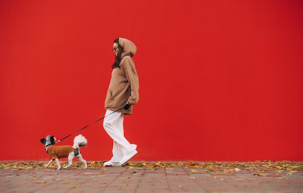 Stylish woman walks with a small dog on a leash on the street on a background of red walls