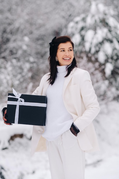 A stylish woman with a white suit with a New Year's gift in her hands in a winter forest A girl in nature in a snowy forest with a gift box