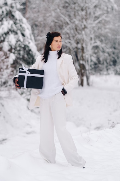 A stylish woman with a white suit with a New Year's gift in her hands in a winter forest A girl in nature in a snowy forest with a gift box