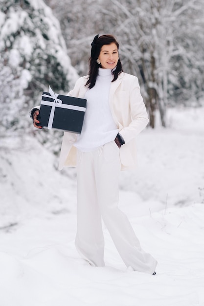 A stylish woman with a white suit with a New Year's gift in her hands in a winter forest A girl in nature in a snowy forest with a gift box