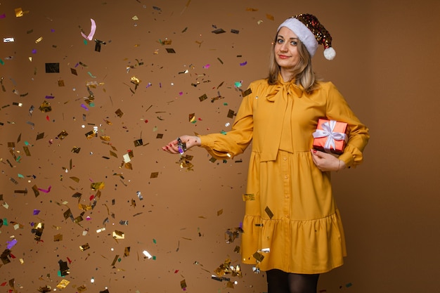 Stylish woman in yellow dress, black shoes and red christmas hat holds a red box with a gift with a lot of confettin around her