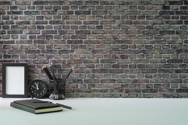 Luogo di lavoro elegante portapenne e taccuino con cornice vuota su un tavolo bianco contro un muro di mattoni con spazio per la copia