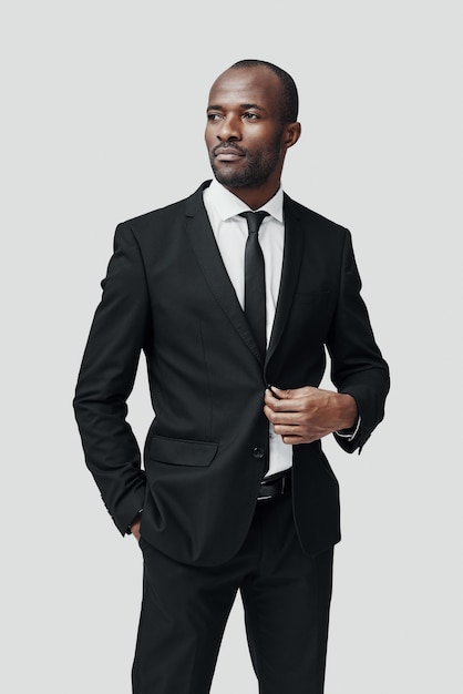 Stylish young African man in formalwear looking away and adjusting jacket while standing against grey wall