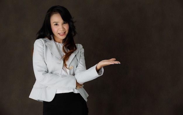 Стильная молодая азиатская бизнес-леди в элегантной одежде улыбается и демонстрирует пустое пространство на сером фоне