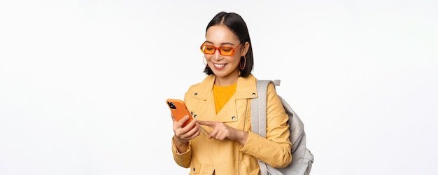 Stylish young asian woman tourist traveller with backpack and smartphone smiling at camera posing against white background