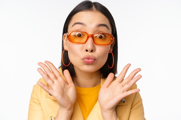 Stylish young asian woman in trendy sunglasses pucker lips silly looking surprised impressed reaction standing over white background