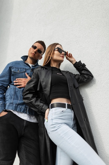 Stylish young beautiful couple in fashionable casual urban clothing with a leather black coat and jeans stand near the wall Fashionable pretty hipster woman wearing cool vintage fashion sunglasses