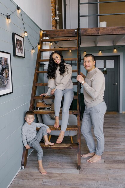 Stylish young beautiful family with a young son pose standing or sitting on a wooden staircase in their country house