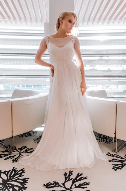 Фото Стильная молодая блондинка позирует в длинном свадебном платье в красивом интерьере днем