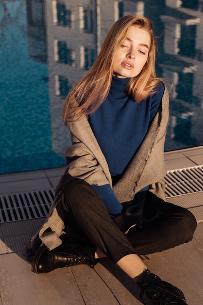 Stylish young blonde girl sits by the blue pool, around her neck a scarf, in the rays of the sun