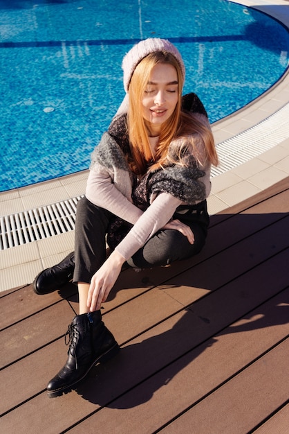 Стильная молодая блондинка модель в розовой шляпе сидит у синего бассейна и улыбается на солнце