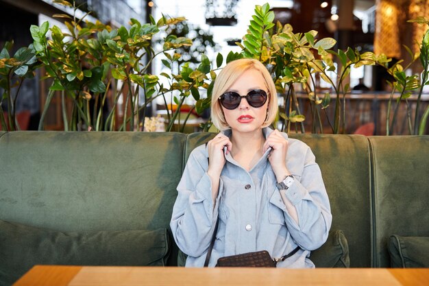 Stylish young blonde in sunglasses sitting at a table in a beautiful restaurant waiting for an order The woman has a break in the working day
