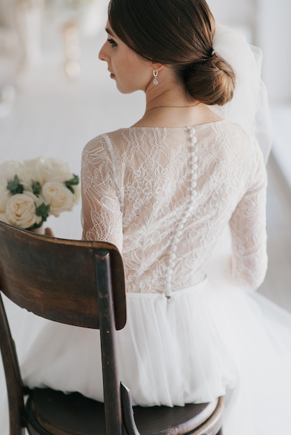 Stylish young bride in a white dress sits on an old wooden chair