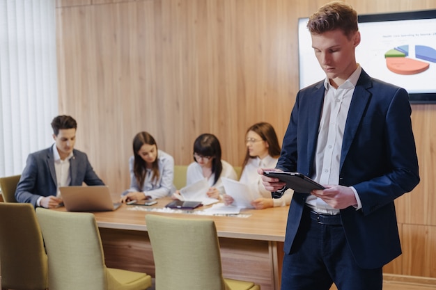 Giovane uomo d'affari alla moda che indossa una giacca e una camicia di un ufficio di lavoro con persone che lavorano con un tablet