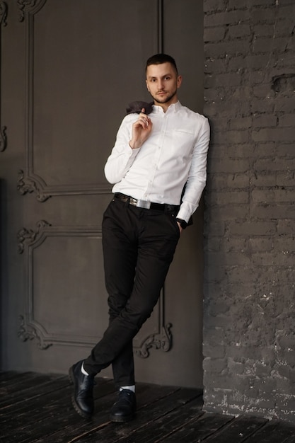 Stylish young businessman in white shirt is holding a jacket on finger standing against brick wall  Vertical photo