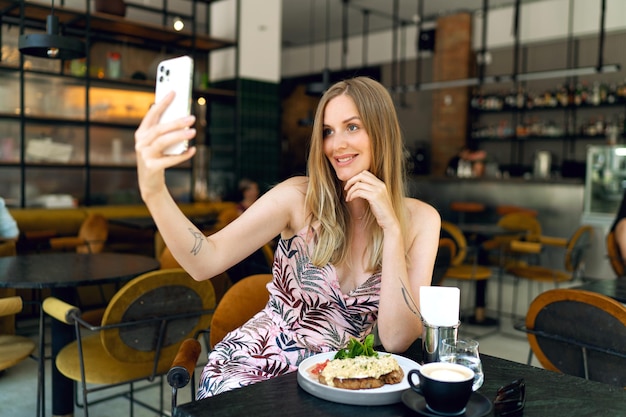 スタイリッシュな若い実業家がブランチカフェで屋内でポーズをとり、彼女のスマートフォン、エレガントな衣装をすすりながら。