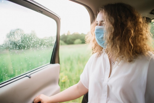 BLの車の後部座席に座りながら窓の外を眺める医療マスクをしたスタイリッシュな若いカーリー女性