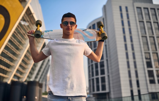 Photo stylish young caucasian american skater holding longboard in hand, in glasses, against the background of urban buildings. the guy put a longboard on his shoulders