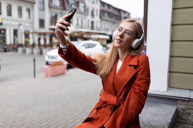 ヘッドフォンでスタイリッシュな若い白人女性が街でセルフィーを作る