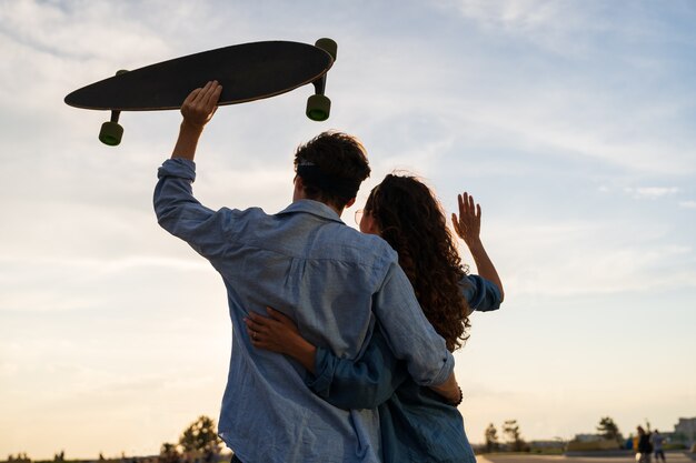 Стильная молодая пара обнимается, глядя на закат, держат скейтборд, счастливая девушка и влюбленный парень наслаждаются свободой